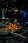 Crop faceless worker in gloves and uniform welding metal details on table near constructions in factory — Stock Photo