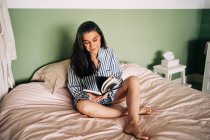 Entspannte reife hispanische Frau in lässiger Kleidung sitzt im Bett und liest interessantes Buch — Stockfoto