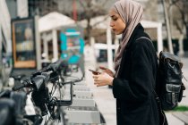 Vista lateral de la mujer musulmana en pañuelo para la cabeza utilizando el sistema de bicicletas compartidas en la ciudad - foto de stock
