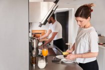 Vista lateral da calma jovem mulher em t-shirt branca preparando café da manhã saudável com abacate enquanto está à mesa na cozinha perto de namorados irreconhecíveis lavar pratos — Fotografia de Stock