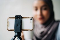 Low angle of cheerful Muslim female in headscarf shooting video on smartphone on tripod for blog while sitting at table in cafe — Stock Photo