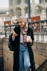 Mulher étnica encantada em hijab e roupas da moda andando lá em cima com bebida takeaway enquanto navega na Internet no smartphone e desfruta de fim de semana na cidade — Fotografia de Stock