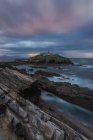 Paisagem de tirar o fôlego da ilha rochosa com farol localizado no oceano perto da costa rochosa em Faro Tapia de Casariego, nas Astúrias, na Espanha, sob céu nublado ao nascer do sol — Fotografia de Stock