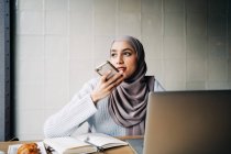 Content ethnic female freelancer in hijab recording audio message on smartphone while sitting at table in cafe and working remotely — Stock Photo