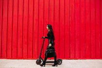 Seitenansicht einer stilvollen Unternehmerin, die mit einem Elektroroller auf dem Bürgersteig in der Stadt gegen eine rote Wand läuft — Stockfoto