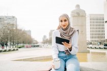 Задоволена мусульманка в хустку сидить з витяжним напоєм біля фонтану в місті і переглядає планшет, дивлячись на камеру. — стокове фото