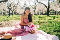 Peaceful Asian woman in pink dress sitting on plaid with blooming flowers in wicker basket in lush garden and looking down — Stock Photo