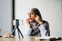 Низький кут веселої мусульманки в хустку знімає відео на смартфоні на тринозі для блогу, сидячи за столом у кафе — стокове фото