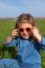 Милая счастливая маленькая девочка в модной одежде и солнцезащитных очках сидит и отдыхает на травянистой лужайке, глядя в камеру — стоковое фото