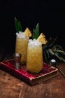 Table en bois avec verres de cocktails jaunes frais avec glaçons et morceaux d'ananas et feuilles près de la cuillère et verre de tir placé sur le livre rouge sur fond noir — Photo de stock