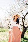 Friedliche nachdenkliche ethnische Frau mit Strohhut und Kleid steht unter blühenden duftenden Blumen auf Ästen im Obstgarten — Stockfoto