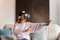Conteúdo jovem do sexo feminino em roupa casual assistindo vídeo usando óculos VR enquanto se senta em um sofá confortável em casa — Fotografia de Stock