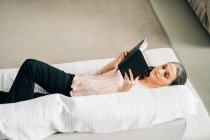 Dall'alto del libro di lettura femminile mentre sdraiato su un comodo divano in soggiorno con pianta verde a casa — Foto stock