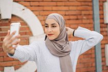 Joven mujer musulmana sonriente en el hiyab tradicional de pie en la calle y tomando uno mismo disparo en el teléfono inteligente - foto de stock