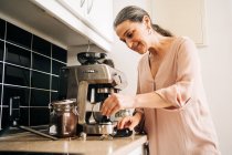 Vista lateral de la hembra de mediana edad concentrada preparando café fresco mientras usa la cafetera moderna en el mostrador de la cocina con teléfono inteligente en el trípode - foto de stock