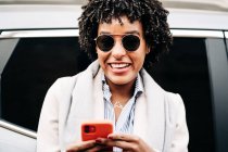 Lachende Afroamerikanerin mit aufgerissenem Mund und stylischer Sonnenbrille mit Handy in der Nähe moderner Fahrzeuge — Stockfoto