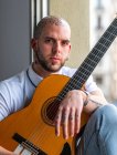 Pensive male with tattooed bald head in casual clothes sitting on windowsill and playing guitar in daytime — Stock Photo