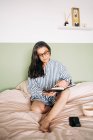 Femme gaie d'âge moyen en chemise rayée souriante et écriture carnet sur lit avec smartphone — Photo de stock
