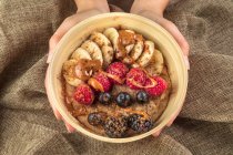 Top view of crop anonymous cook showing bowl with banana slices and assorted fresh berries with caramel sauce for breakfast — Stock Photo