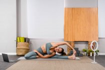 Full body of flexible calm young lady in sportswear stretching spine in Revolved Head to Knee pose while practicing yoga in studio near smartphone on tripod and laptop — Stock Photo