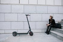Vista laterale di stili imprenditore donna in berretto seduto sulle scale in città vicino scooter elettrico moderno — Foto stock