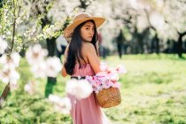 Вид ззаду на жінку в сукні та солом'яному капелюсі, що стоїть з кошиком у квітучому саду, дивлячись на плече до камери — стокове фото