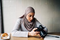 Focused Muslim female photographer sitting at table and looking through photos on professional camera while working remotely in cafe — Stock Photo