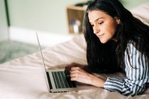 Вид сфокусированной зрелой, самозанятой латиноамериканки с длинными темными волосами в повседневной одежде, лежащей на кровати и печатающей на ноутбуке во время онлайн-работы дома — стоковое фото