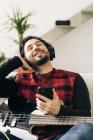 Adulto barbudo músico masculino con bajo guitarra en los auriculares de mensajes de texto en el teléfono celular en la sala de estar - foto de stock