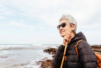 Vue latérale de trekker femme âgée souriante dans des lunettes de soleil avec les cheveux gris regardant loin contre l'océan orageux — Photo de stock