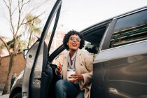 Glückliche Afroamerikanerin in modischer Sonnenbrille mit Thermobecher und Smartphone, während sie lächelt und silbernes Prestigeauto zurücklässt — Stockfoto