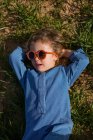 Зверху дівчина в модному одязі і сонцезахисних окулярах тримає руки за голову і розслабляється на трав'яному газоні — стокове фото