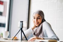 Baixo ângulo de fêmea muçulmana alegre em vÃ deo de tiro de lenço de cabeça no smartphone no tripé para blog enquanto se senta Ã mesa no café — Fotografia de Stock