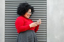 Вид збоку пухнастої усміхненої жінки в яскравому текстовому повідомленні на мобільний телефон, спираючись на ребристу стіну в місті — стокове фото