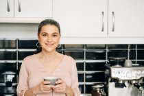 Allegro femmina di mezza età con tazza di bevanda calda in piedi al bancone della cucina con armadi bianchi e macchina da caffè moderna a casa — Foto stock
