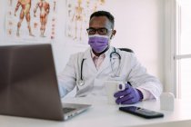 Médico masculino afroamericano concentrado irreconocible en bata médica y máscara beber café y trabajar en el ordenador portátil en la clínica moderna - foto de stock