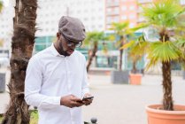 Homem afro-americano na moda em pé na rua com palmeiras e mensagens nas mídias sociais via telefone celular — Fotografia de Stock