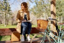 Молода вдумлива жінка робить нотатки в планувальнику на дерев'яній лавці біля велосипеда в парку вдень — стокове фото