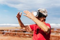 Anonymous elderly female traveler experiencing virtual reality in goggles on sea shore under cloudy sky — Stock Photo