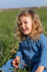 Lindo niño feliz sentado en el campo verde en el día soleado mirando hacia otro lado y jugando con la hierba en verano - foto de stock