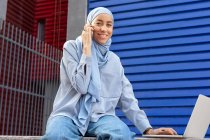 Lächelnde Muslimin mit Kopftuch und Netbook spricht auf dem Handy, während sie in die Kamera in der Stadt schaut — Stockfoto