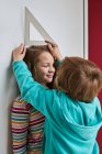 Irmão ajudando irmã com medir sua altura com régua e lápis perto da parede — Fotografia de Stock