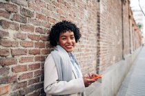 Delighted African American female with curly hair standing against brick wall and browsing mobile phone — Stock Photo