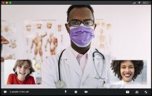 Unrecognizable concentrated African American male doctor in medical robe and mask looking at camera while communicating with positive multiethnic female and boy during video call — Stock Photo