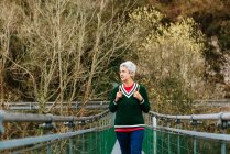 Elderly female hiker in casual clothes strolling on suspension bridge while looking away in daytime — Stock Photo