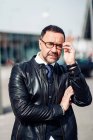 Serious middle aged ethnic male executive in eyewear and leather jacket touching arm while looking at camera in town — Stock Photo