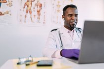 Médico masculino afro-americano concentrado em robe médico e fones de ouvido TWS trabalhando em laptop enquanto sentado à mesa na clínica moderna — Fotografia de Stock