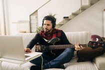 Adulto músico masculino em fones de ouvido tocando baixo contra netbook no sofá na sala de estar — Fotografia de Stock