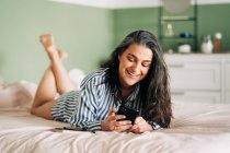 Positive hispanische Frau mittleren Alters mit langen dunklen Haaren in lässiger Kleidung lächelt, während sie auf dem Handy zu Hause im Bett liegt — Stockfoto