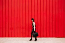 Вид збоку на жіночого підприємця в модному одязі, що йде вулицею на фоні червоної стіни і дивиться на камеру — стокове фото